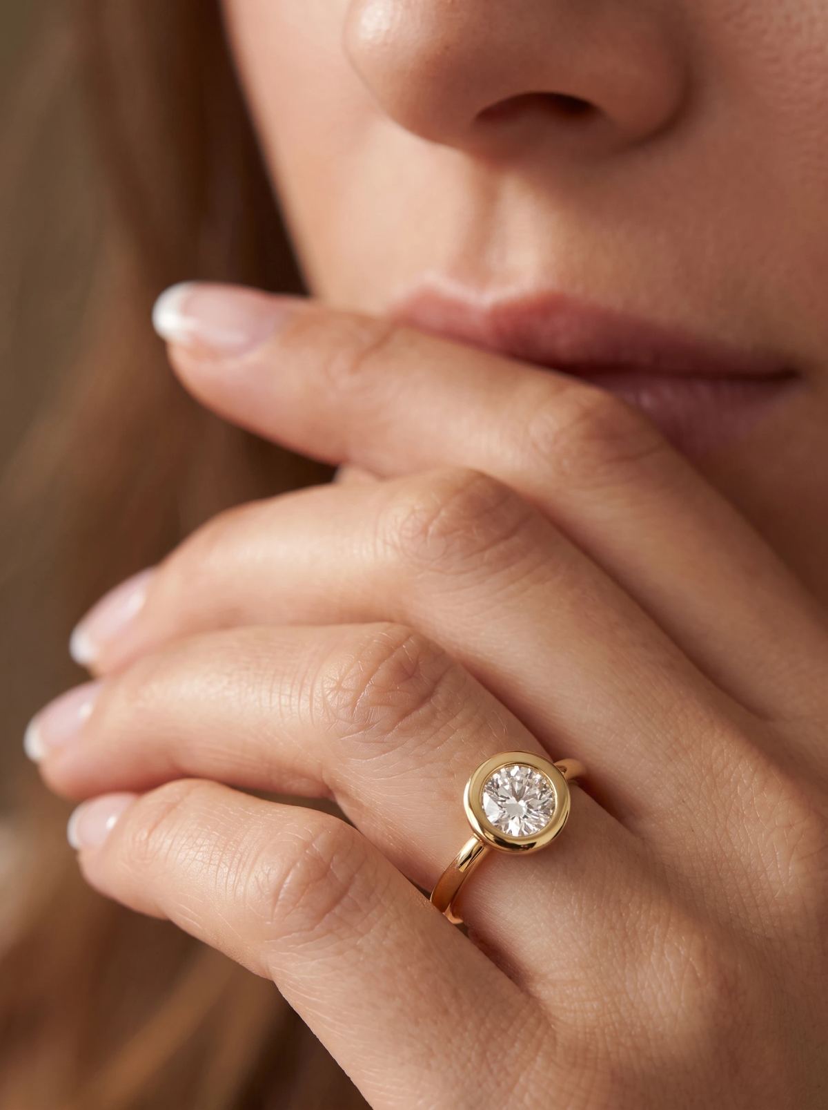 Bague de fiançailles en serti clos en or jaune portée à la main, doigts posés contre les lèvres