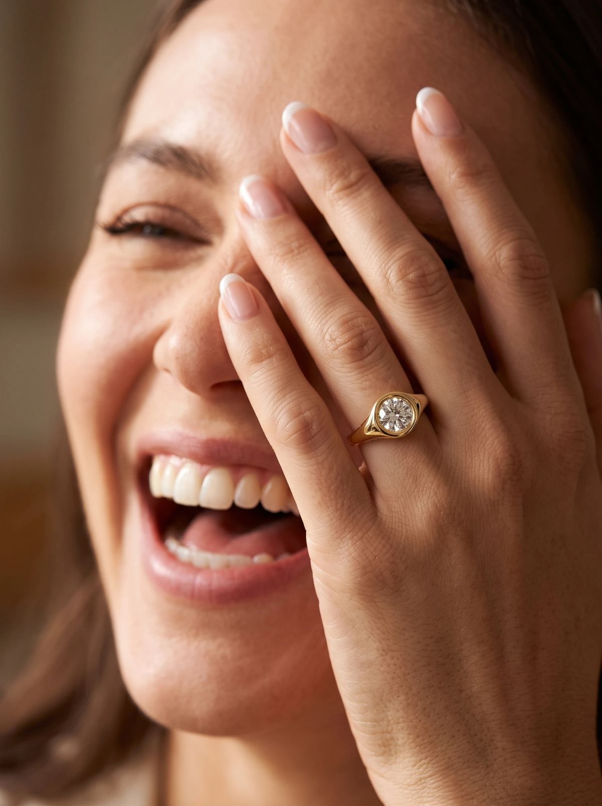 Chevalière de fiançailles en or jaune à diamant rond portée par une femme riant, la main devant l'œil