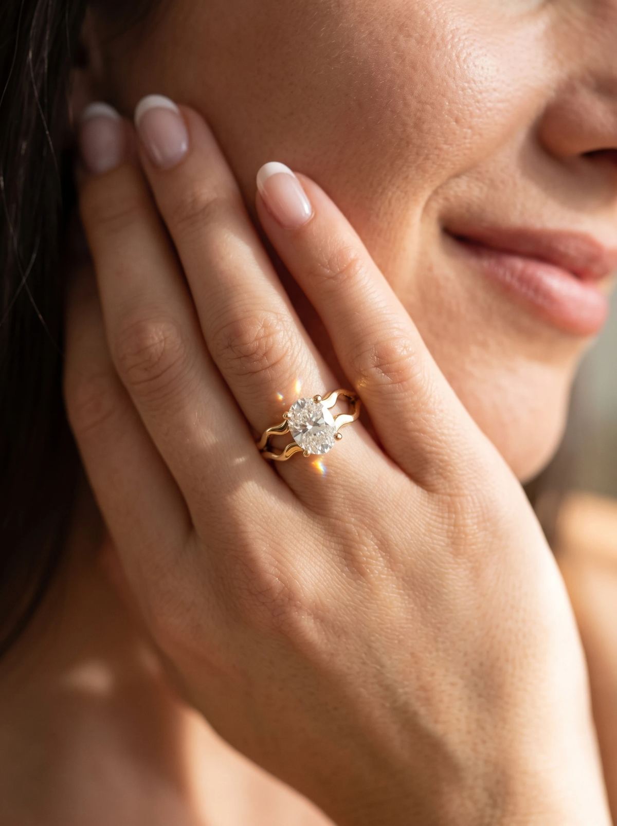 Bague de fiançailles ovale à anneau divisé en or jaune portée sur une main posée contre la joue