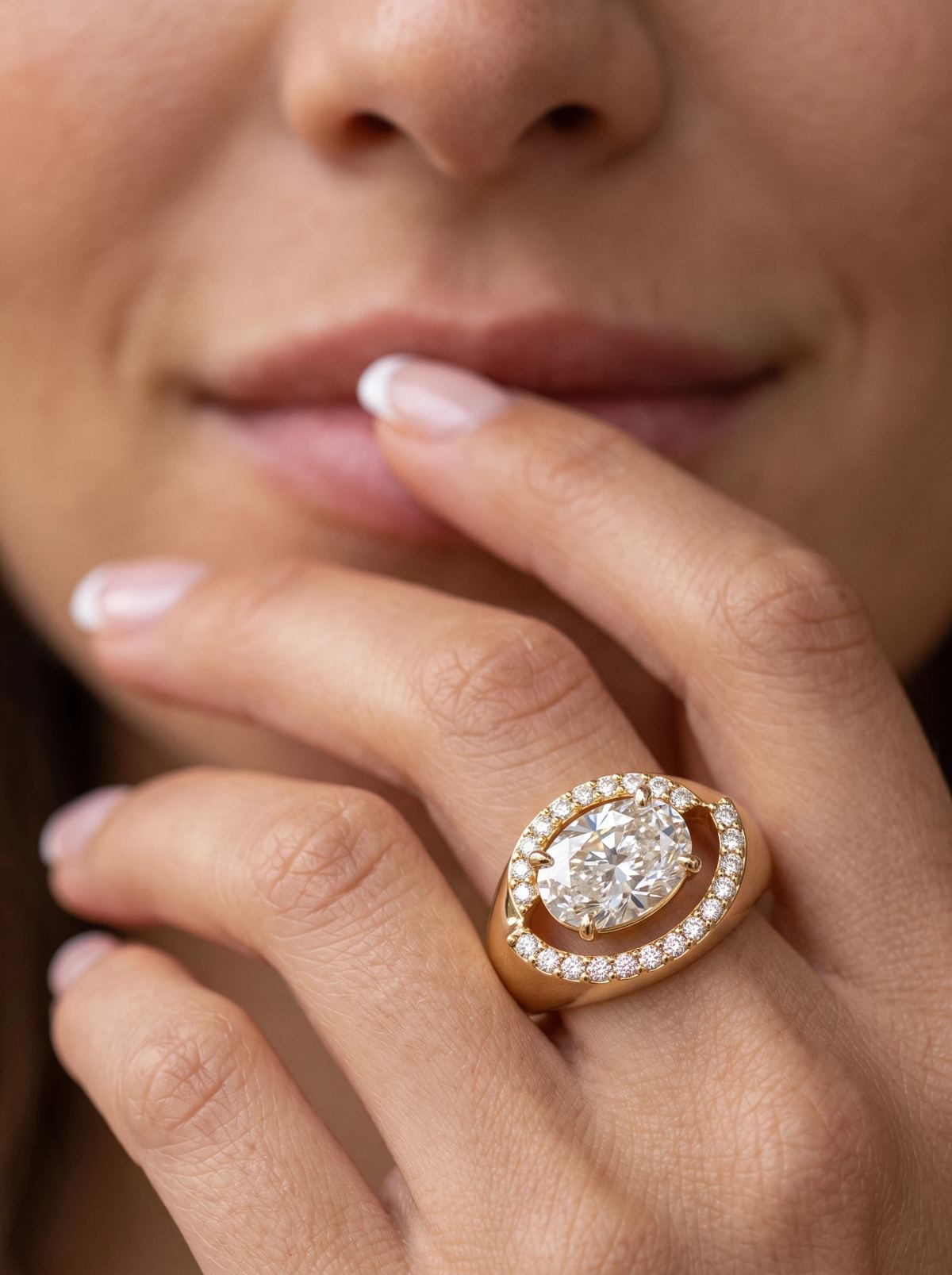 Bague de fiançailles ovale est-ouest en or jaune portée à la main près des lèvres, halo décalé face à l'objectif