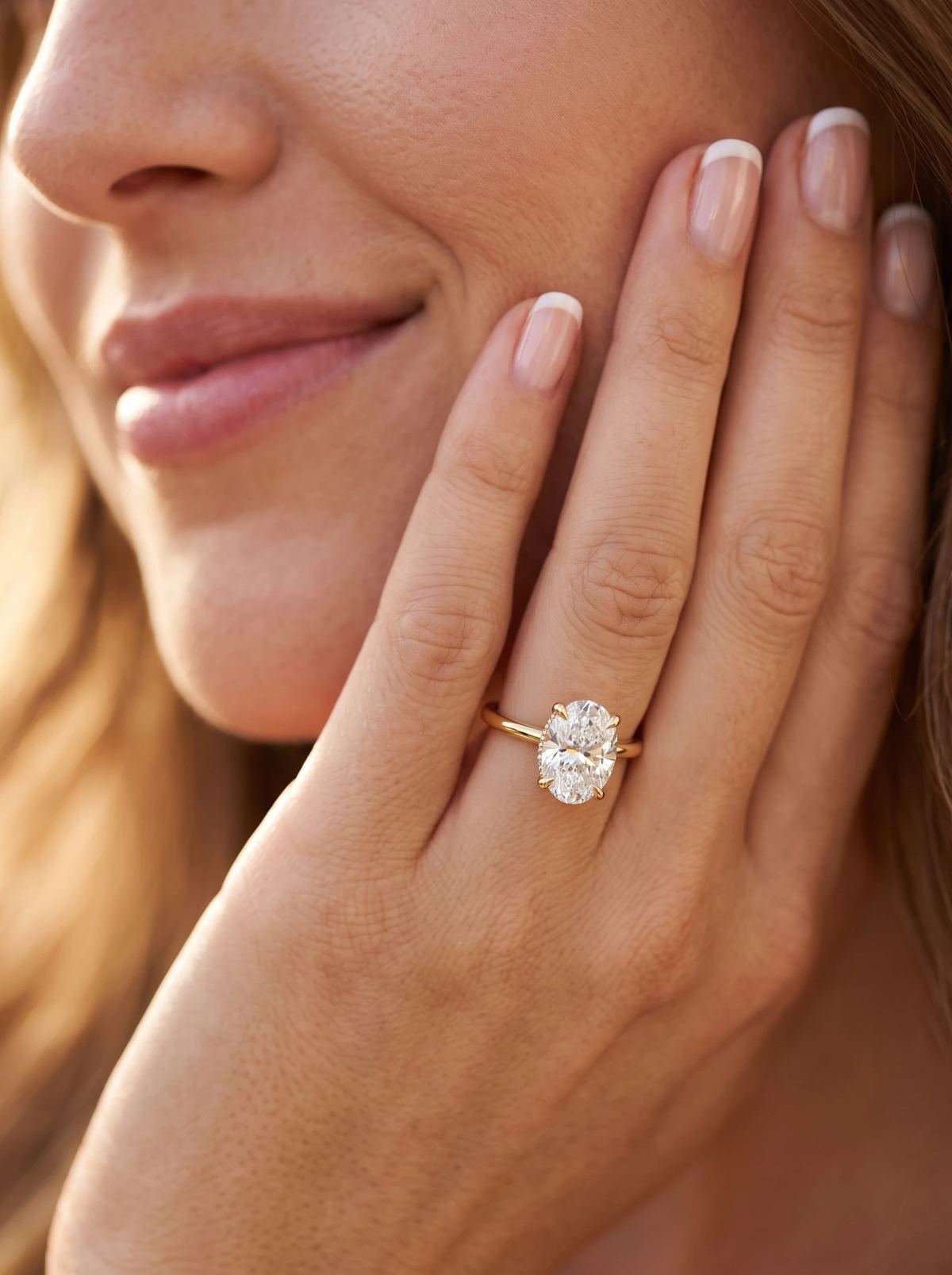 Bague de fiançailles ovale à halo caché en or jaune portée sur une main posée contre la joue