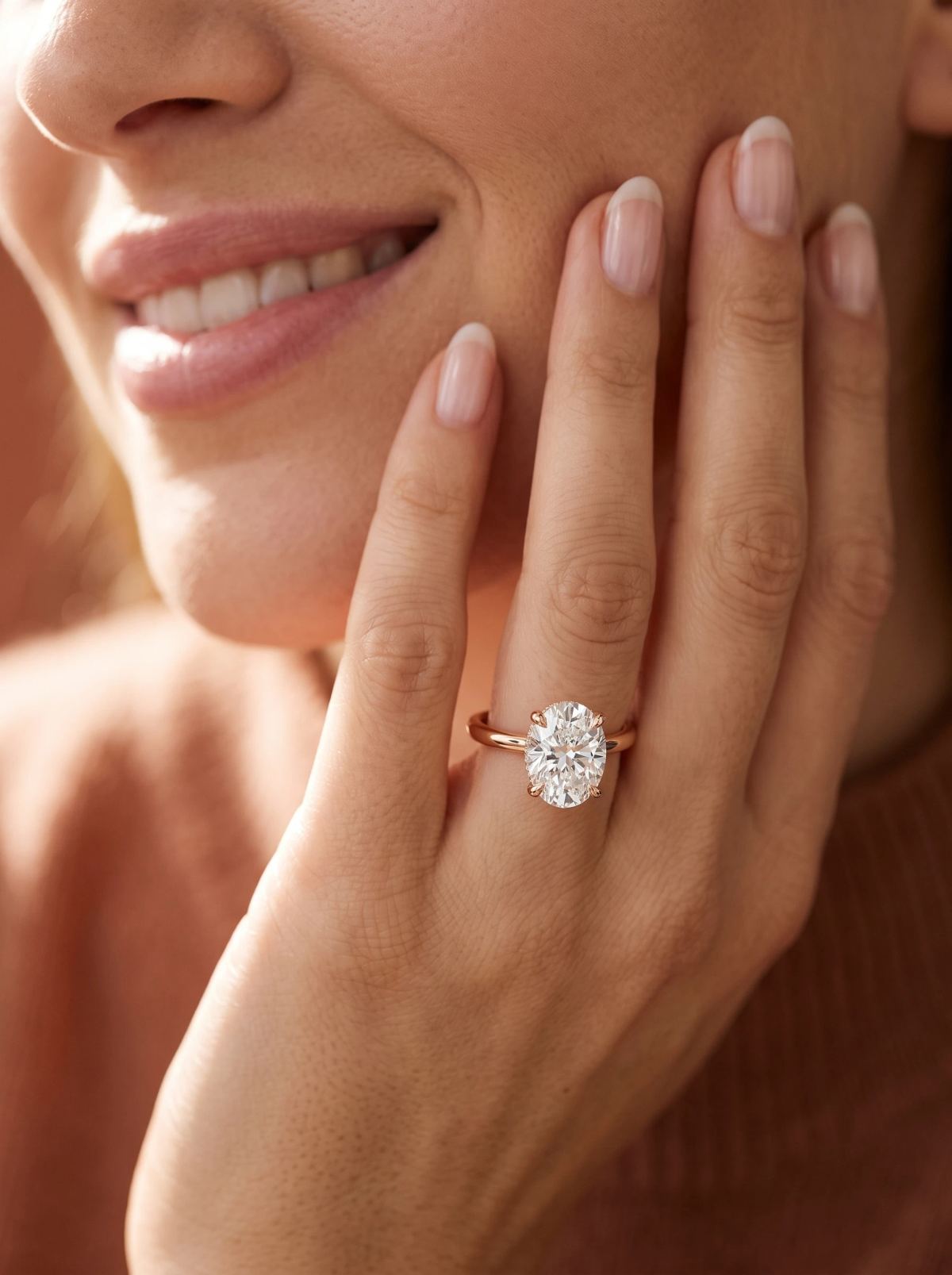 Solitaire ovale en or rose porté à la main, posée contre la joue d'une femme souriante